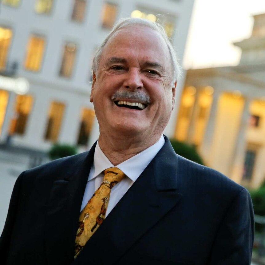 John Cleese in a suit and moustache standing outside the Brandenburg Gate