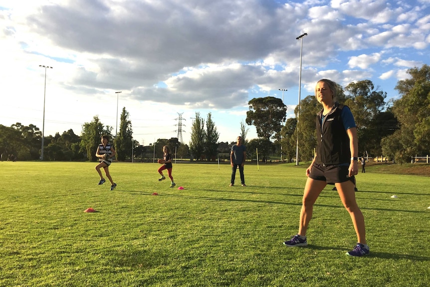 Brooke Patterson stands to the left as young footballers run around markers on a football oval.