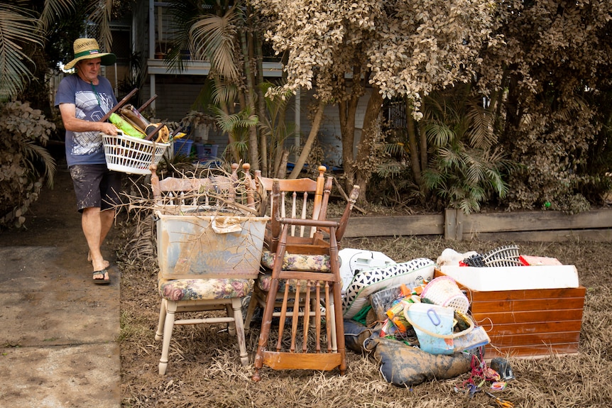 A man carries a basket of ruined items to a pile of damaged goods left on the curb.