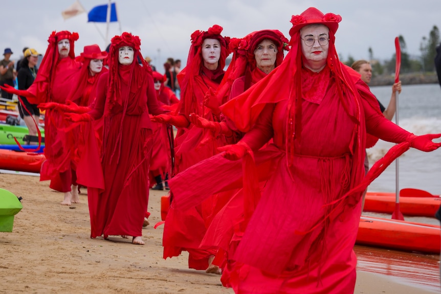 Six women and a man lined up behind each other wearing bright red costumes and painted white faces.