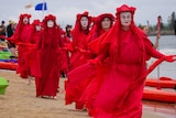 Six women and a man lined up behind each other wearing bright red costumes and painted white faces.