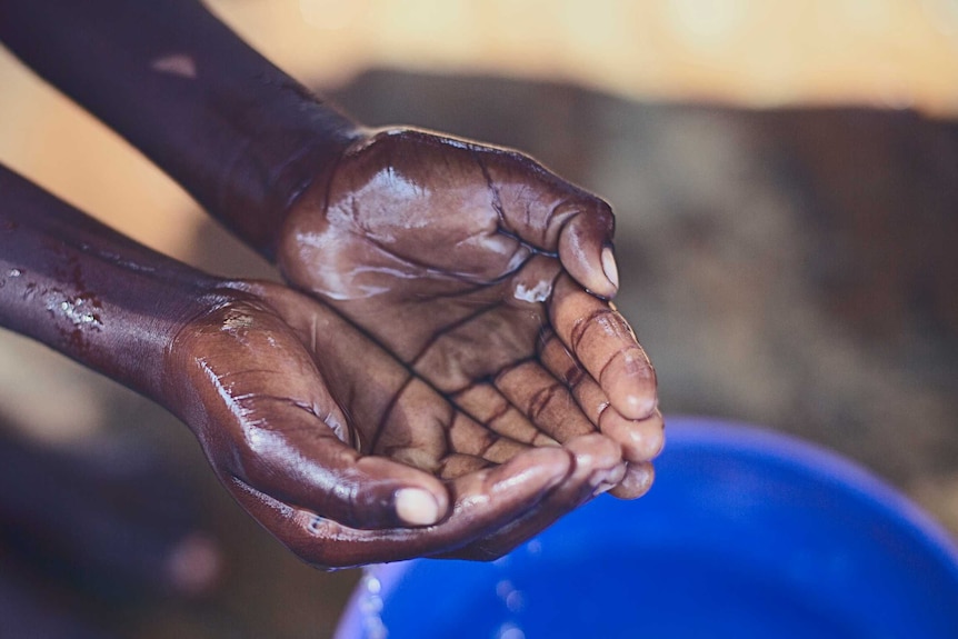 The hands of a child holding water they have picked up from a blue tub that sits underneath.