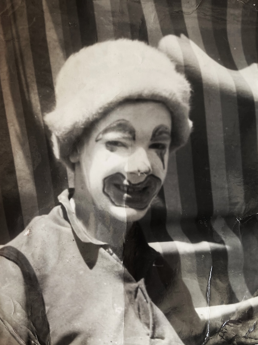 A black and white photo of a young person in clown make up with striped circus behind