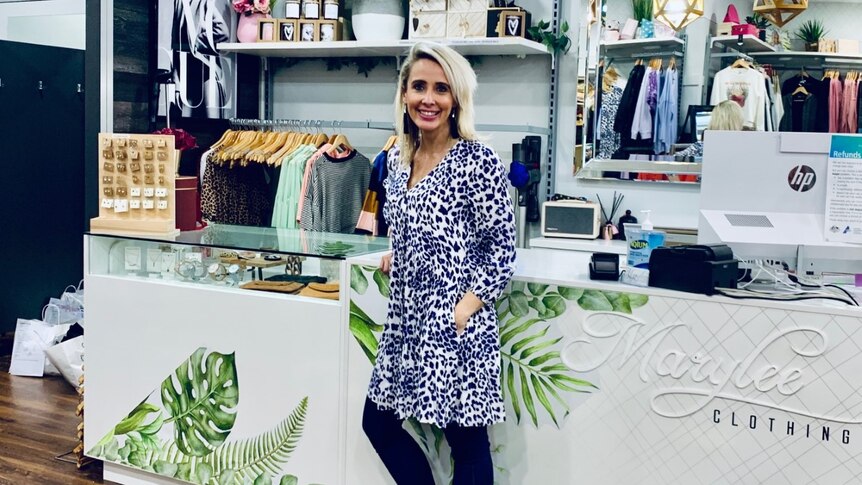 A young blonde haired woman stands smiling in front of a counter in a clothing store, with tops hanging behind her.