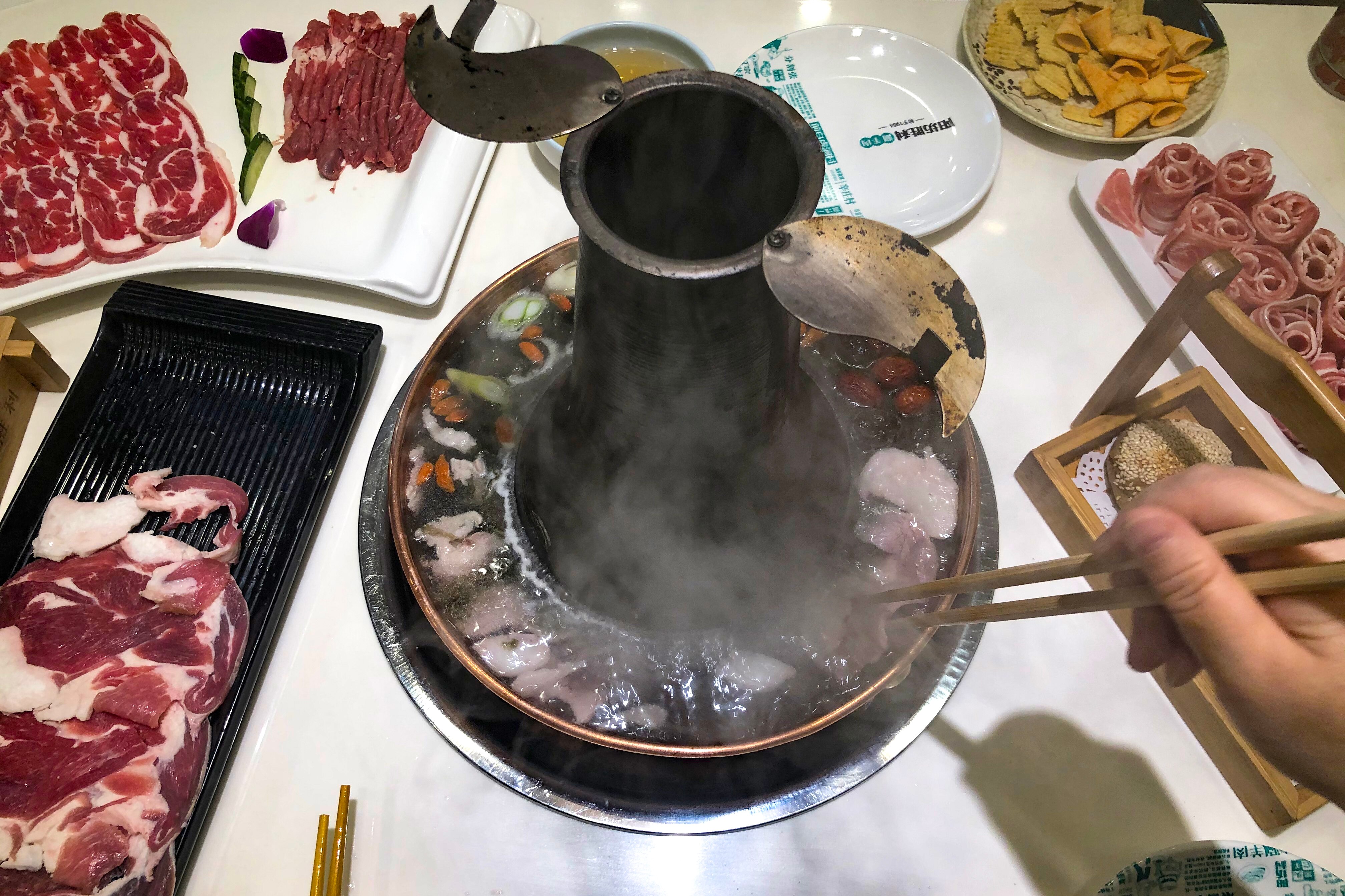 A hand holding chopsticks dunks thinly sliced raw red meat into a steaming Chinese hot pot.