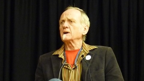 A man sits besides a drum, speaking to a crowd.