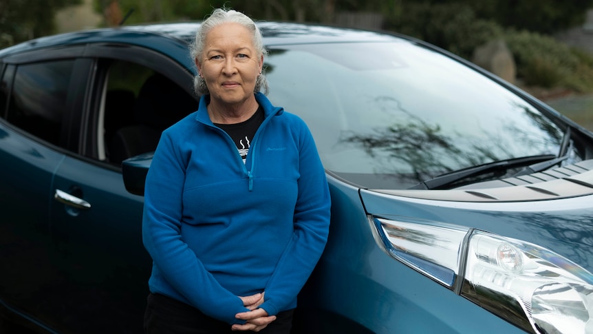  A woman with grey hair and a blue jumper stands in front of a blue car