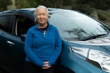  A woman with grey hair and a blue jumper stands in front of a blue car