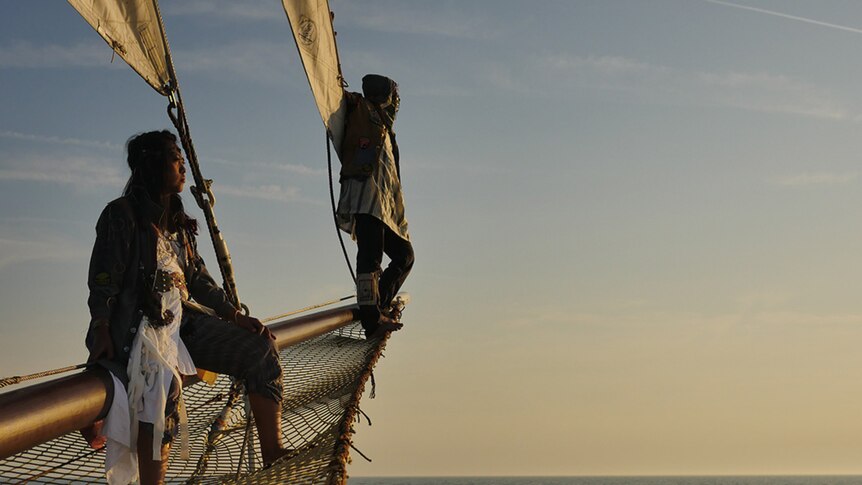 Nova and Grey on the netted bow of their ship against a warm sunset over sea