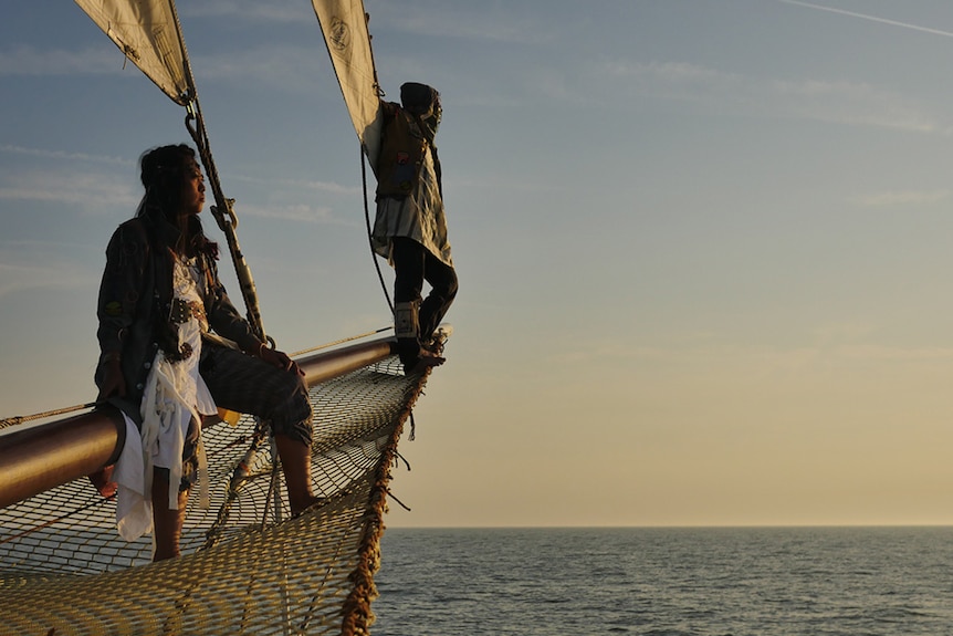 Nova and Grey on the netted bow of their ship against a warm sunset over sea