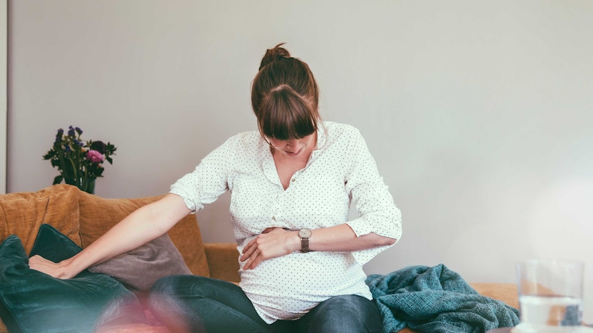A pregnant woman holds her stomach while sitting on a couch