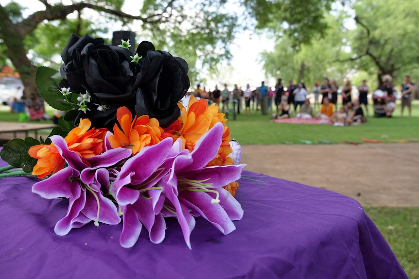A photo of black, orange and purple flowers on what looks like a casket, a group of people blurred in the background.