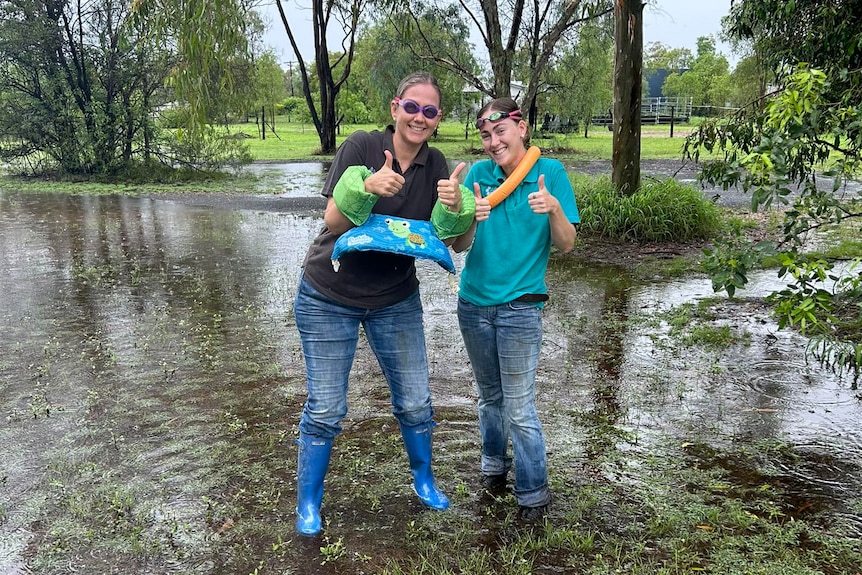 Two women wearing gum boots and snorkelling gear give thumbs up while standing on wet ground with trees around them