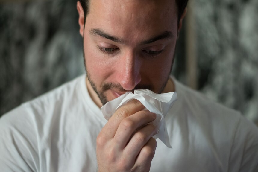 A man with dark hair wearing a white t-shirt wipes his nose with a tissue