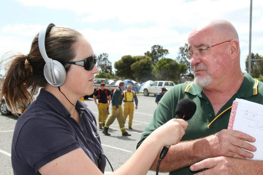 Reporter with headphones on holding microphone and interviewing a man holding papers with firefighters in carpark in background.