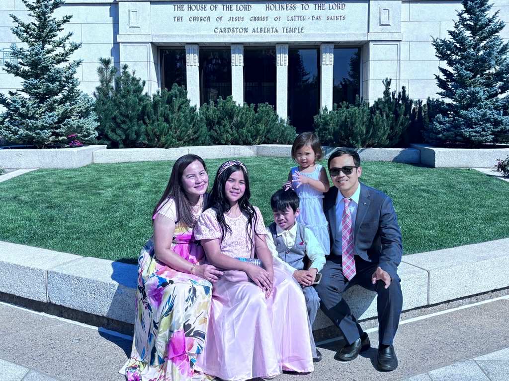 Una familia de cinco miembros (un hombre, una mujer, dos niñas y un niño pequeño) está sentada en un arcén pavimentado alrededor de un trozo de hierba. 