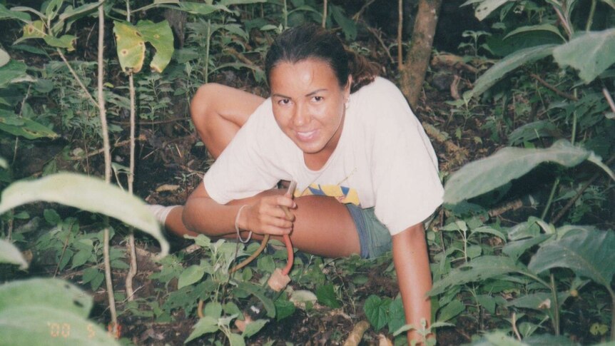 A woman on the ground in a forested area holds scientific equipment.