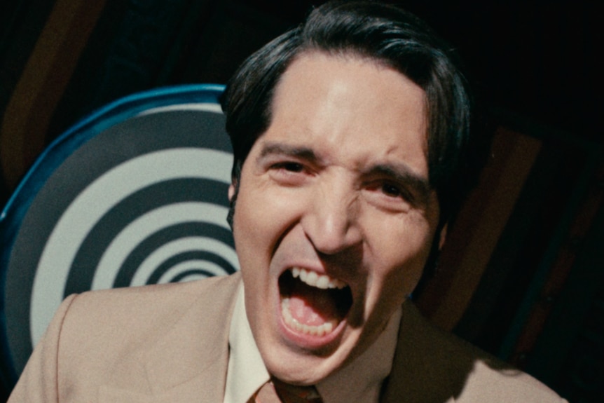 A tilted close up of a man screaming, with a spiralling hypnotising board behind him.