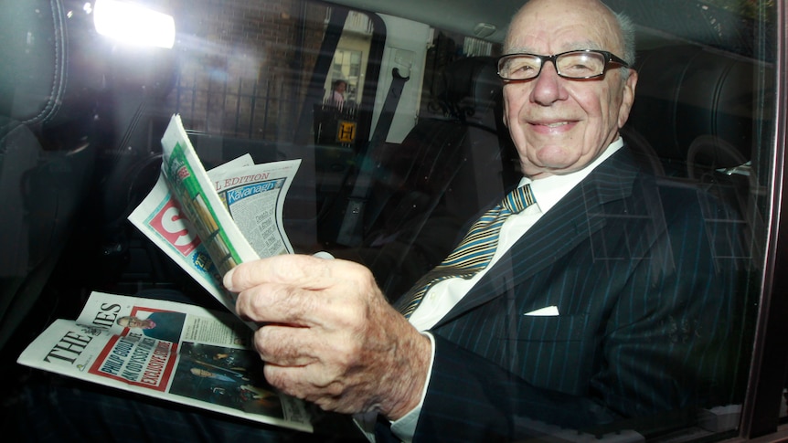 Rupert Murdoch, an older man with glasses, reads newspapers in the back of a car.