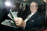 Rupert Murdoch, an older man with glasses, reads newspapers in the back of a car.