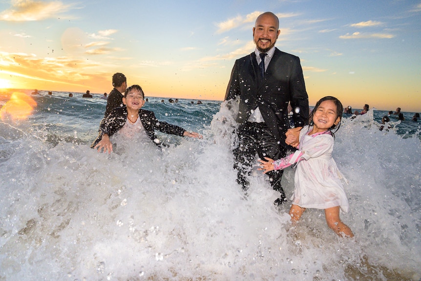 Two young children and a father play in the ocean in formal outfits.