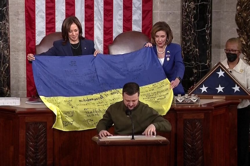 A man in combat fatigues addresses the US Congress as Kamala Harris and Nancy Pelosi hold a signed Ukrainian flag behind him.