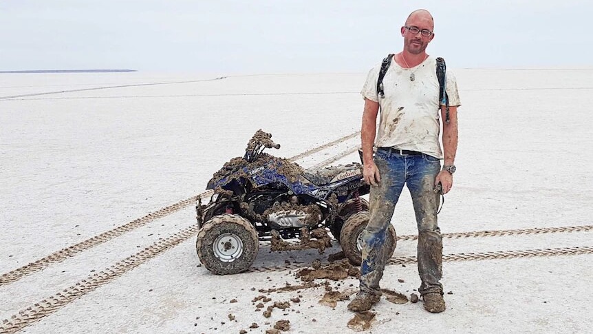 Man wearing a muddy white t-shirt and jeans, standing in front of a quad bike in desert.