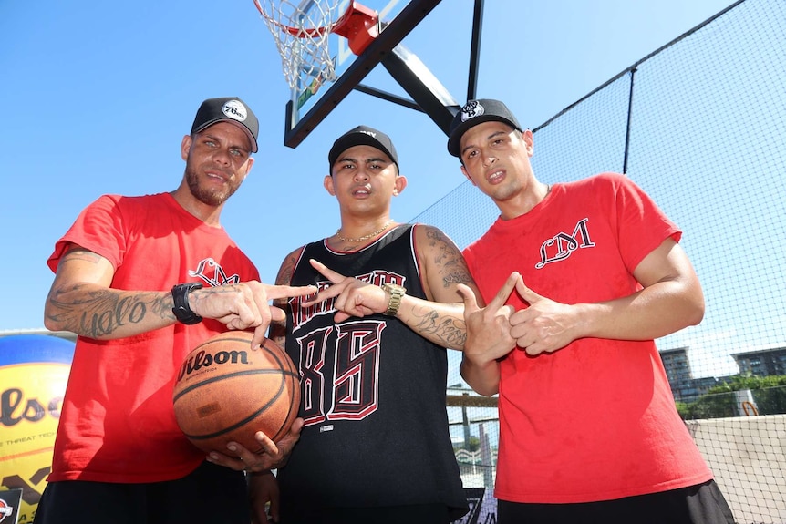 James Ryan, John Micaran and Jaydee Martin create hand gestures in front of a basketball ring.