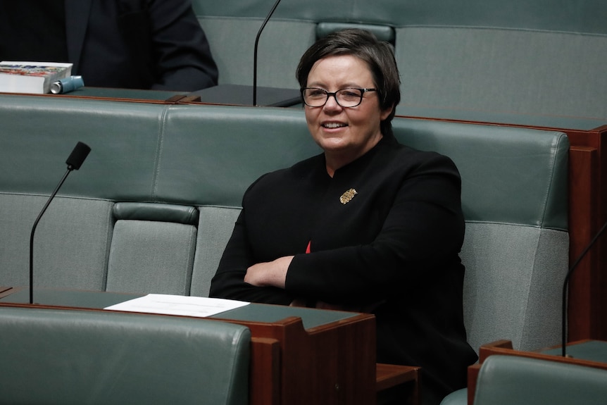 A woman with short brown hair and glasses sits on a grey blue seat in front of a microphone