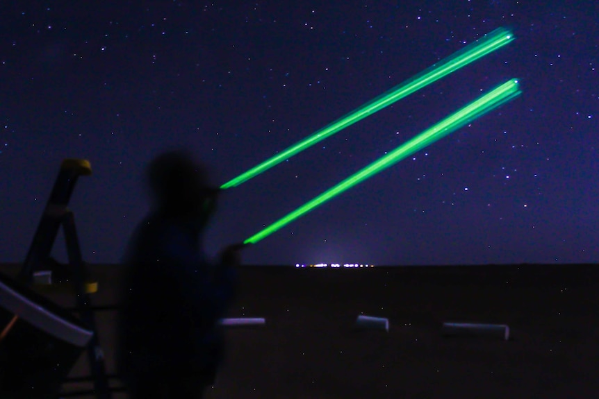 A man in shadow shines green lasers across a blue starry sky. Burketown's lights can be seen, dotted in the background.