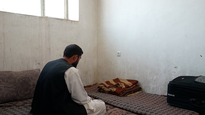 A man sits on the floor of a bare room, his back to the camera. The white walls are stained. Rugs are on the floor.