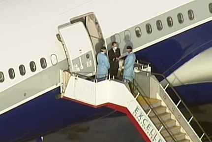 A plane sits on the tarmac as two people stand at the top of the departure stairs wearing masks.
