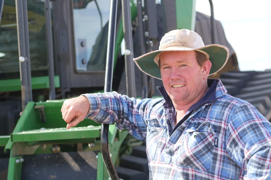 A man leans on a tractor looking at the camera. He is wearing a blue shirt. 