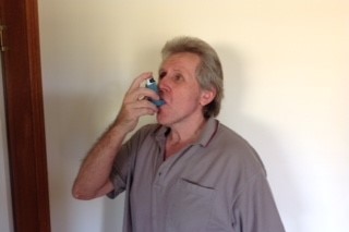 An older man with light hair and tee-shirt uses  an inhaler indoors.