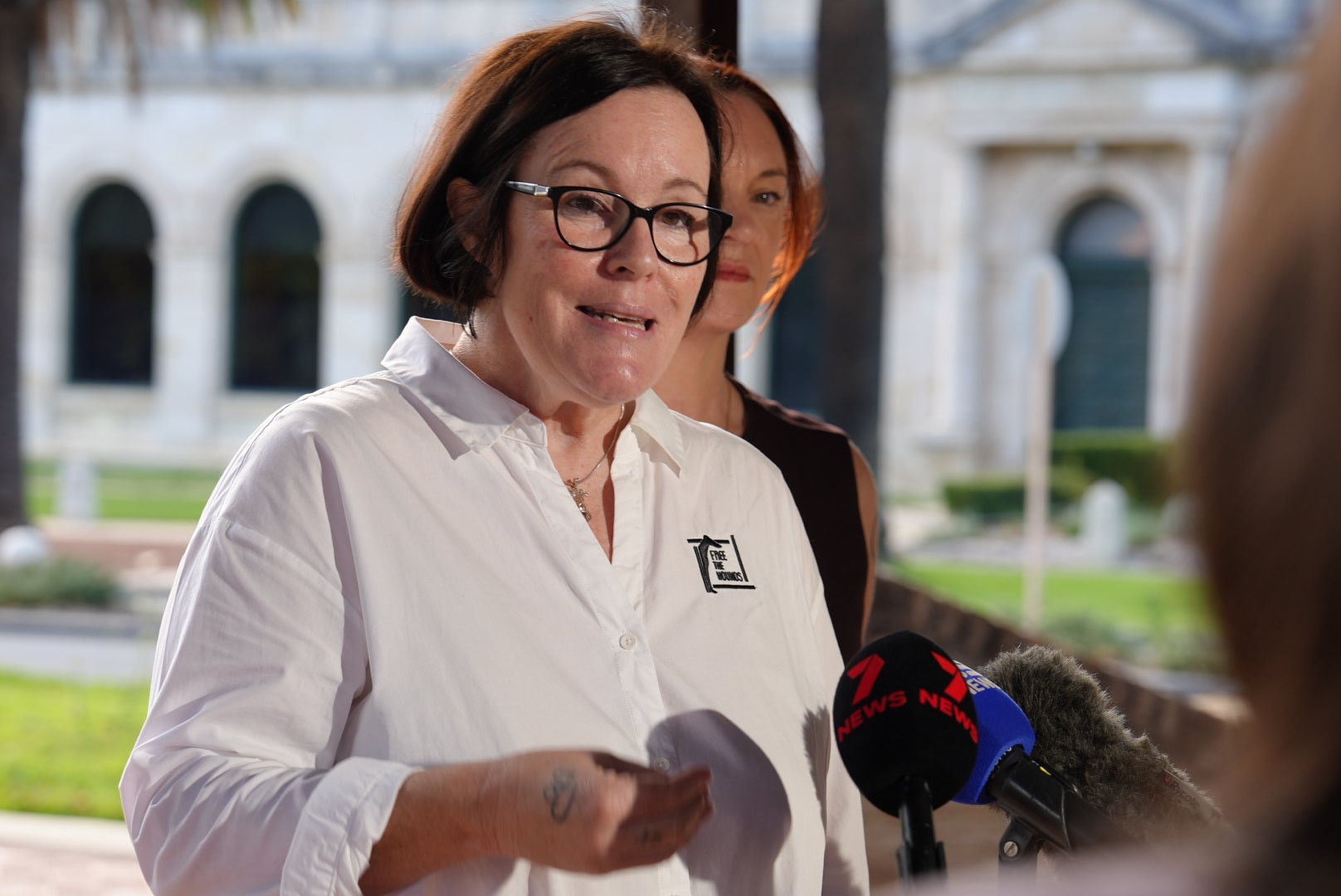 Free the Hounds president Melissa Harrison stands speaking during a media conference outside, wearing a white shirt.