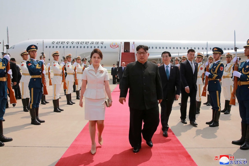 Kim Jong-un and his wife Ri Sol Ju, dressed in a cream skirt suit, walk down a red carpet with a plane in the background