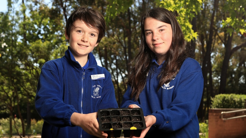 Levi and Holly wear blue jumpers and hold a black seedling pot outside while smiling.