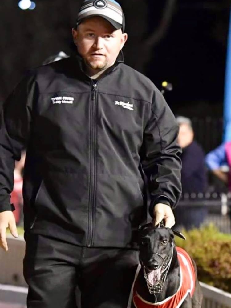 A man in a black jacket and cap walks with a black greyhound.