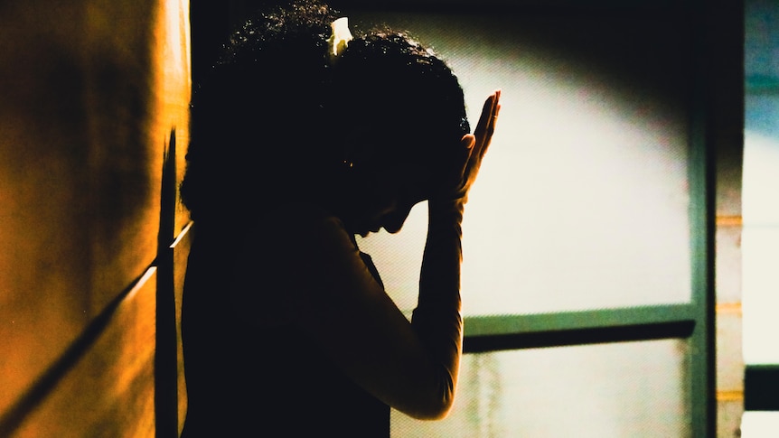 A woman holds her palm to her forehead as she stands in a corridor, silhouetted.