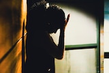 A woman holds her palm to her forehead as she stands in a corridor, silhouetted.