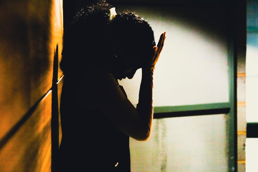 A woman holds her palm to her forehead as she stands in a corridor, silhouetted.