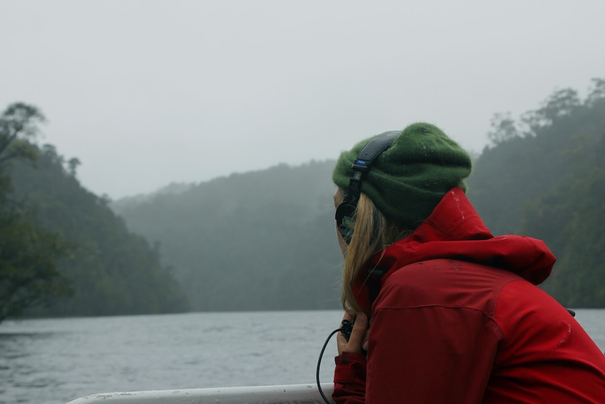 A woman wearing headphones and a red jacket looks at the misty river and forest behind a boat.