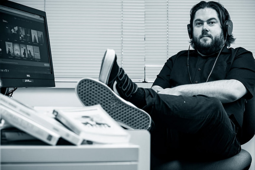 Black-and-white photo of a man with a beard sitting in an office smiling with his feet resting on the desk.