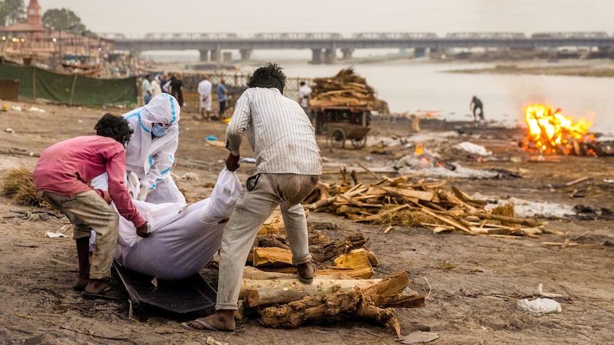 A man in full PPE and two men in regular clothes move a body wrapped in cloth on the banks of a river