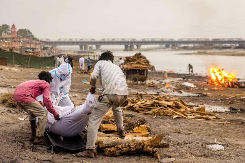 A man in full PPE and two men in regular clothes move a body wrapped in cloth on the banks of a river