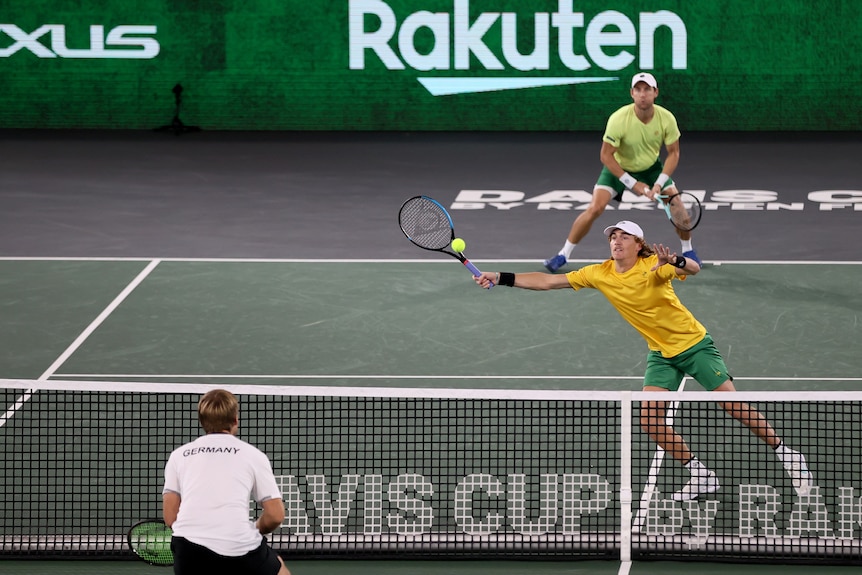 An Australian Davis Cup doubles player reaches for a volley at the net, while his partner remains on the baseline.