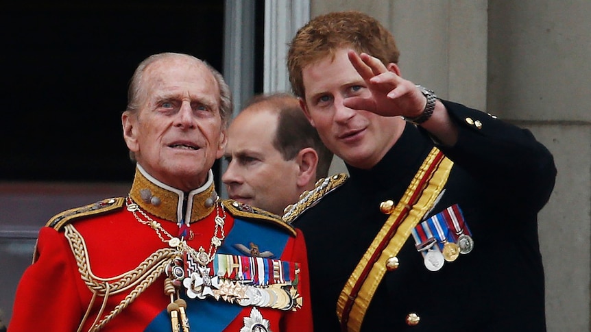 A man appears in a red jacket decorated with medals on the left next to a younger man in a black jacket with some medals too.