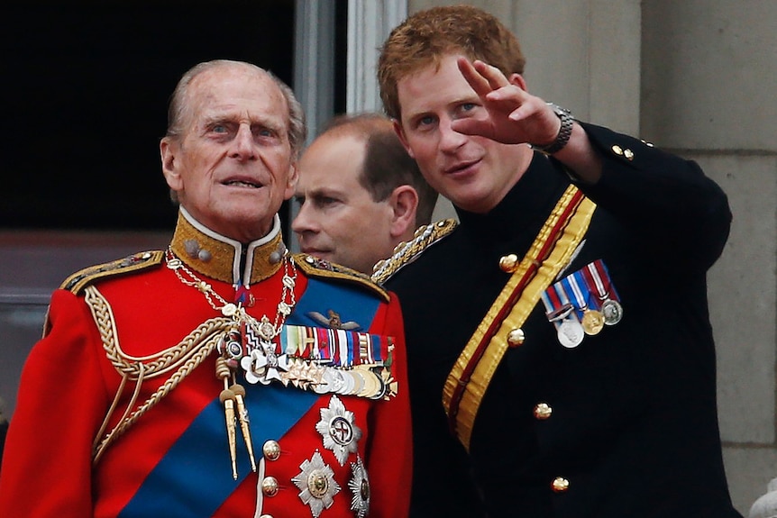 A man appears in a red jacket decorated with medals on the left next to a younger man in a black jacket with some medals too.