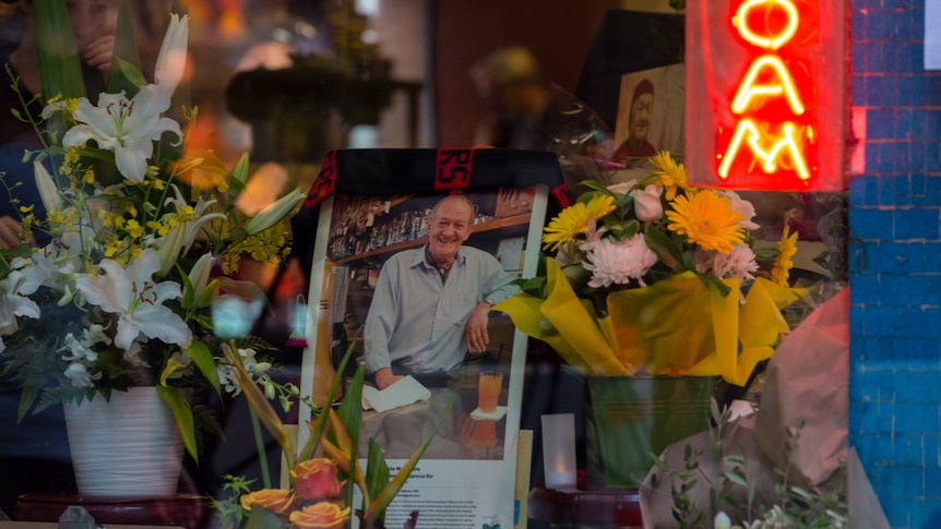 An AFL Bombers flag is draped over a photograph of Sisto Malaspina, surrounded by flowers in the window of the cafe.
