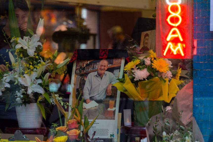 An AFL Bombers flag is draped over a photograph of Sisto Malaspina, surrounded by flowers in the window of the cafe.